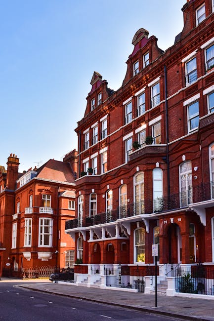 A street scene showing a multi-storey red brick building with large windows and decorative architectural details, situated on a residential street in West Kensington. The building features white window frames, small balconies with black wrought iron railings, and a white entrance canopy supported by decorative brackets. The pavement in front is clear, with a parking restriction sign posted near the curb. The scene is illuminated by natural daylight under a bright blue sky. This image relates to house removals and relocation services provided by Removals West Kensington, highlighting the type of properties that may require professional furniture transport, packing, and loading during home relocation processes.