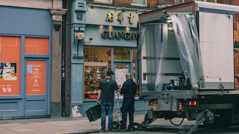 Two movers from Removals West Kensington are preparing a home relocation outside a shop storefront with Chinese signage in West Kensington. One mover is carrying a green plastic crate, while the other is standing next to a hand truck with a cardboard box. The scene includes a professional moving truck parked nearby, with its side door open and covered with a transparent plastic sheet to protect the interior. The background shows the shop's blue facade, large windows displaying various items, and some signage in Chinese characters above the entrance. The pavement is clean, and the area appears well-lit, indicating daytime conditions. This image captures the process of furniture transport and packing in preparation for a house or business move, exploring typical loading or unloading activities involved in professional removals services.