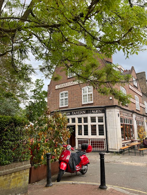 A street scene showing a multi-storey red brick building with large windows and decorative architectural details, situated on a residential street in West Kensington. The building features white window frames, small balconies with black wrought iron railings, and a white entrance canopy supported by decorative brackets. The pavement in front is clear, with a parking restriction sign posted near the curb. The scene is illuminated by natural daylight under a bright blue sky. This image relates to house removals and relocation services provided by Removals West Kensington, highlighting the type of properties that may require professional furniture transport, packing, and loading during home relocation processes.