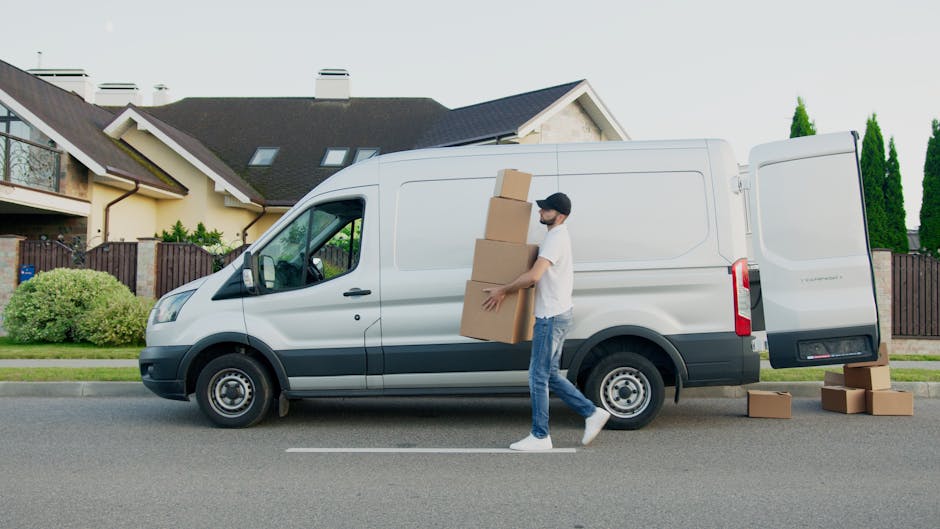 A man wearing a white polo shirt and sunglasses is standing outside a residential house on a suburban street during sunset, seen through the open rear doors of a white moving van. The van's interior i