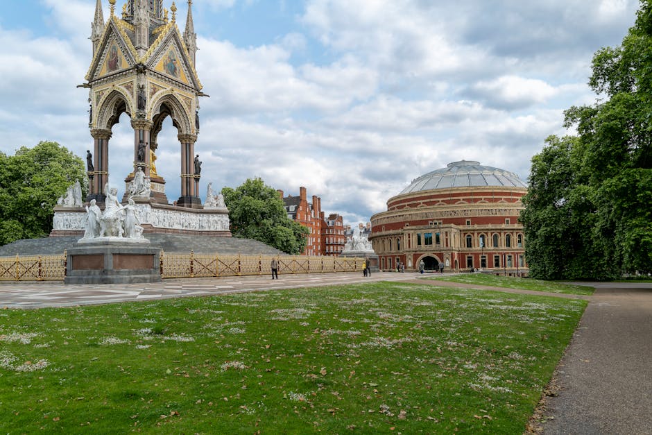 A wide outdoor view of a public park area featuring a large ornate, gilded monument with statues and decorative elements, situated on a raised platform surrounded by a wooden fence. To the right, there is a historic circular building with a domed roof and red brick façade, positioned near a paved walkway and lush green trees. The sky is partly cloudy with patches of blue, and a few people are visible walking in the distance. This scene captures an urban park space, which may be relevant to house removals or moving logistics in the West Kensington area, where [COMPANY_NAME] provides professional removals and furniture transport services.