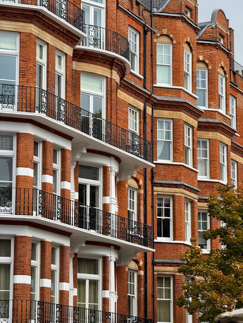 A multi-storey residential building with a traditional red brick exterior, featuring white decorative stone accents around the windows and columns. Several balconies with black wrought iron railings are visible on each floor, extending from the building's façade. The image shows the building's exterior from a street-level perspective during daylight, with a small tree partially visible on the right side, its leaves indicating autumn. The scene is clear, with natural lighting highlighting the architectural details. This setting is typical of urban house removals or furniture transport preparations, relevant to professional relocation services such as those offered by Removals West Kensington, especially near West Kensington station.
